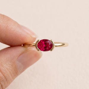 May include: A gold ring featuring a vibrant oval-cut red gemstone set in a tension-style mounting. The band is smooth and polished, holding the faceted stone securely between two curved metal prongs that frame the sides of the oval gem.