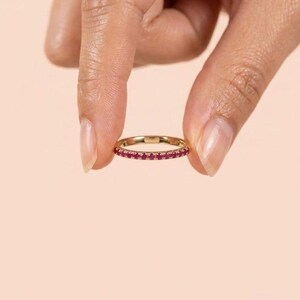 Pode incluir: Um anel de banda fina em ouro com uma fila cont&iacute;nua de pequenas pedras preciosas redondas de cor rosa vibrante cravadas ao longo da circunfer&ecirc;ncia exterior. O metal tem um acabamento polido e as pedras est&atilde;o espa&ccedil;adas uniformemente.