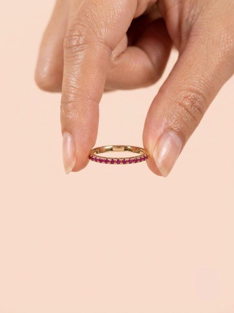 Pode incluir: Um anel de banda fina em ouro com uma fila cont&iacute;nua de pequenas pedras preciosas redondas de cor rosa vibrante cravadas ao longo da circunfer&ecirc;ncia exterior. O metal tem um acabamento polido e as pedras est&atilde;o espa&ccedil;adas uniformemente.