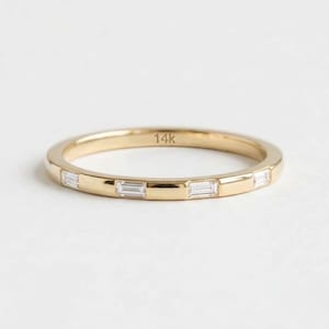 May include: A thin yellow gold band ring featuring four evenly spaced baguette-cut clear stones set horizontally along the outer surface. The interior of the band is clearly engraved with the text 14k, indicating the gold purity of the metal.