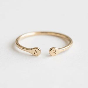 May include: A gold-toned open ring featuring two rounded ends, each engraved with a single capital letter. The left end displays the letter A, and the right end displays the letter R. The band has a smooth, polished finish throughout its circular shape.