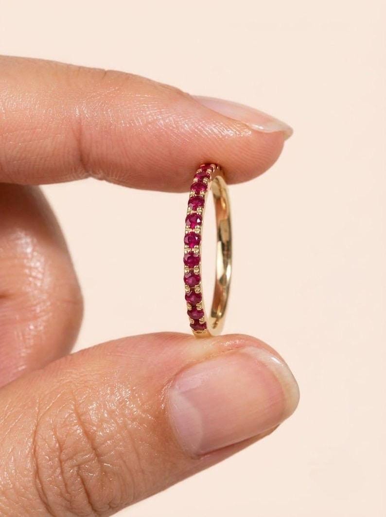Pode incluir: Um anel de banda fina em ouro com uma fila cont&iacute;nua de pequenos rubis redondos de um vermelho vibrante cravados ao longo da circunfer&ecirc;ncia exterior. O anel apresenta um acabamento em metal polido e um alinhamento delicado de gemas.