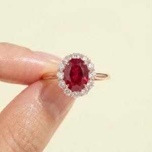 May include: A rose gold ring featuring a central oval-cut deep red ruby gemstone surrounded by a halo of small, round, brilliant-cut white diamonds. The band is thin and polished, showcasing a classic prong setting for both the center stone and the surrounding diamond accents.