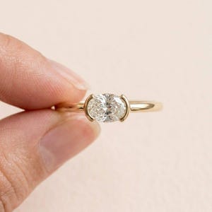 May include: A gold ring featuring an oval-cut diamond. The diamond is set in a bezel setting, and the band is thin and polished. The ring is held by a hand against a soft pink background. A classic and elegant piece of jewelry.