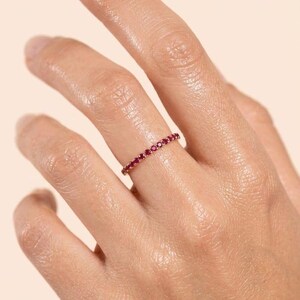 Pode incluir: Um anel fino e delicado com uma banda cont&iacute;nua de pequenas pedras preciosas redondas de um vermelho vibrante, cravadas num metal polido de tom dourado. O anel envolve o dedo, exibindo uma fila uniforme de pedras vermelhas cintilantes.