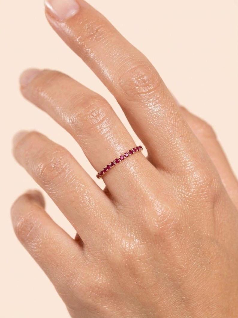 Pode incluir: Um anel fino e delicado com uma banda cont&iacute;nua de pequenas pedras preciosas redondas de um vermelho vibrante, cravadas num metal polido de tom dourado. O anel envolve o dedo, exibindo uma fila uniforme de pedras vermelhas cintilantes.
