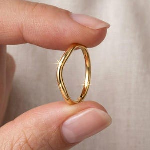 Può includere: Un anello in oro lucido caratterizzato da un design sottile e ondulato lungo la fascia. La superficie metallica è liscia e riflettente, cattura la luce creando un brillante riflesso metallico sulla forma curva del gioiello.
