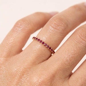 Pode incluir: Um anel de banda fina em ouro com uma fila cont&iacute;nua de pequenos rubis redondos de um vermelho vibrante, cravados em garras partilhadas. O anel &eacute; usado num dedo, destacando o trabalho delicado do metal e o tamanho consistente das pedras.