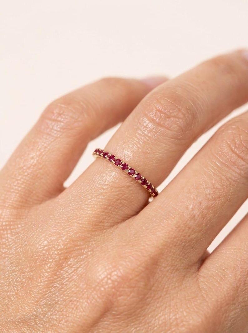 Pode incluir: Um anel de banda fina em ouro com uma fila cont&iacute;nua de pequenos rubis redondos de um vermelho vibrante, cravados em garras partilhadas. O anel &eacute; usado num dedo, destacando o trabalho delicado do metal e o tamanho consistente das pedras.