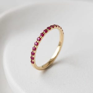 May include: A thin gold band ring featuring a row of small, round, vibrant red rubies set along the top half of the circumference. The metal has a polished finish, and the gemstones are evenly spaced and securely held in place by small prongs.