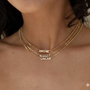 Puede incluir: Tres collares dorados con colgantes con nombres. El collar superior dice "ARCHIE", el del medio "KAILEY" y el inferior "OSCAR". Los collares se llevan alrededor del cuello.