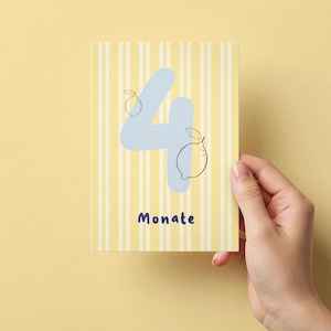 Può includere: Una carta con sfondo a righe gialle e bianche. Il numero 4 è azzurro chiaro con illustrazioni di limoni. La parola "Monate" è scritta in blu scuro. La carta è tenuta in mano su uno sfondo giallo.