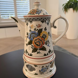 Op de afbeelding: Een witte keramische theepot met bijpassende theelichthouder, voorzien van handgeschilderde bloemmotieven in oranje, blauw, geel en groen. De theepot heeft een deksel, een gebogen handvat en een tuit, en de voet heeft uitgesneden openingen.
