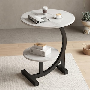 Pode incluir: Uma mesa lateral moderna de dois níveis, com tampo branco com efeito mármore e estrutura de metal preta. A mesa tem um suporte curvo e um tampo circular. Uma xícara de café, um jornal e um copo descartável estão no nível superior.