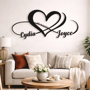 Peut inclure: Décoration murale en métal noir représentant un cœur et un symbole infini. Les noms "Lydia" et "Joyce" sont affichés, avec la date "EST 06.08.2019" à l'intérieur du cœur. L'œuvre est au-dessus d'un canapé avec des coussins.