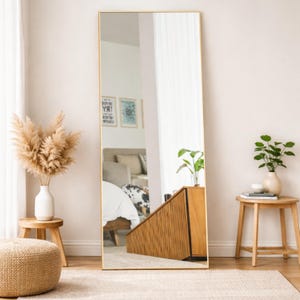 May include: A full-length rectangular mirror with a thin gold frame. The mirror is leaning against a wall in a room with neutral tones. The room is decorated with plants, a wooden cabinet, and a small stool.