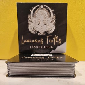 May include: A black oracle deck with gold lettering reading "Luminous Truths". The box displays a white illustration of a woman with a halo. The deck is stacked on top of the box.