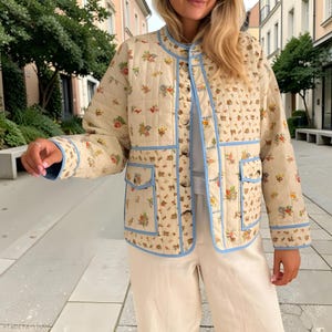 May include: Cream-colored quilted jacket with a floral pattern in red, yellow, and blue. The jacket has a round collar, light blue trim, and two front pockets. The jacket is buttoned up and appears to be made of a soft, lightweight material.
