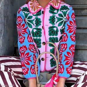 Peut inclure: Veste matelassée colorée avec un motif floral rouge, vert et bleu sur fond rose. La veste a un col arrondi et une fermeture boutonnée. Les manches sont bleues avec des motifs floraux rouges. Un sac à main beige et un pantalon rayé sont également visibles.