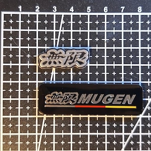 May include: Two rectangular automotive emblems on a grid mat. The top emblem features silver Japanese kanji characters on a black background. The bottom emblem displays silver Japanese kanji, the text MUGEN, and a red, yellow, and black horizontal stripe.