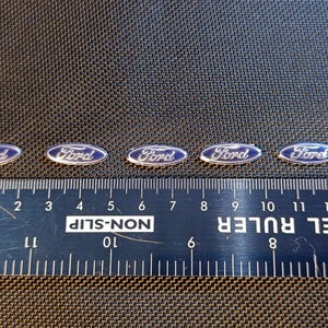 May include: Five oval Ford badges with a blue background and silver lettering are shown. A ruler with both centimetres and inches is placed below the badges, indicating their size. The badges are on a dark textured surface.