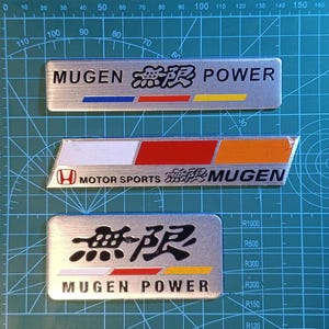 May include: Three rectangular metallic automotive emblems. The top emblem reads MUGEN POWER with Japanese characters and a blue, red, and yellow stripe. The middle emblem features a Honda logo, text MOTOR SPORTS MUGEN, and a white, red, and orange stripe. The bottom emblem displays Japanese characters above MUGEN POWER and a white, red, and yellow stripe.