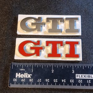 Peut inclure: Deux emblèmes rectangulaires GTI, l'un avec des lettres argentées et l'autre avec des lettres rouges, placés au-dessus d'une règle noire Helix montrant une largeur d'environ 10 cm pour chaque emblème, avec des lettres en relief sur fond blanc.