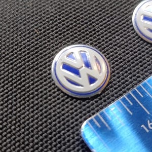 Könnte beinhalten: Zwei silber-blaue Volkswagen-Emblem-Pins. Die Pins haben eine kreisförmige Form mit einem blauen VW-Logo in der Mitte, umgeben von einem silbernen Rand. Ein blaues Lineal mit weißen Markierungen befindet sich im Bild.