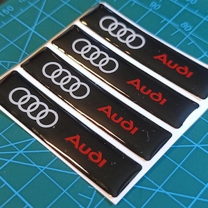 Può includere: Un set di quattro adesivi rettangolari neri con il logo Audi a quattro anelli bianco e il nome del marchio Audi in rosso. Ogni adesivo ha una finitura lucida e in rilievo, disposti in diagonale su un foglio di supporto bianco.