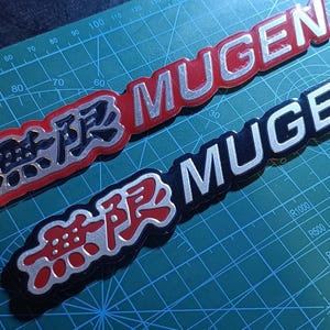 Puede incluir: Dos emblemas Mugen que presentan los caracteres kanji japoneses para Mugen seguidos del texto MUGEN en letras plateadas. Un emblema tiene un fondo rojo y el otro un fondo negro, ambos con bordes plateados, sobre una alfombrilla de corte verde.