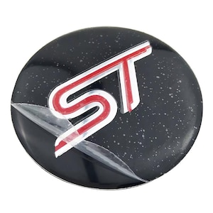 Puede incluir: Un emblema ST negro y plateado con un acento rojo. El emblema es circular y tiene una película protectora parcialmente despegada. Las letras "ST" se muestran de forma destacada en una fuente estilizada.