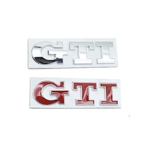 Puede incluir: Dos emblemas automotrices rectangulares con las letras GTI en una fuente de bloque en negrita. El emblema superior tiene un acabado cromado pulido, mientras que el inferior presenta letras rojas con un borde cromado sobre una base plateada.