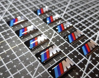 Puede incluir: Una colección de pegatinas rectangulares con el logotipo M, que presenta un diseño de rayas diagonales en azul, morado y rojo sobre un fondo negro. Cada pegatina tiene un acabado brillante y muestra la letra M en una fuente metálica plateada.