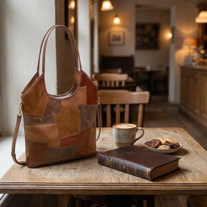 May include: A brown leather tote bag with a patchwork design sits on a wooden table. The bag has a shoulder strap and a handle. A cup of coffee, a plate of cookies, and a book are also on the table.