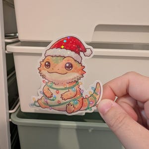 May include: A holographic sticker featuring a cartoon bearded dragon with orange and yellow scales. The lizard wears a red and white Santa hat and is wrapped in a string of colourful Christmas lights. The design has a glossy, reflective finish.