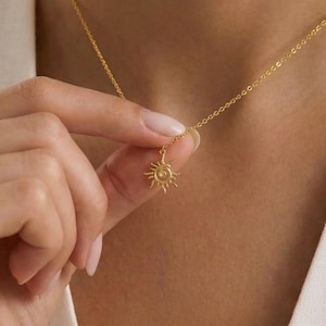 Könnte beinhalten: Eine goldfarbene Halskette mit einem Sonnenanhänger. Der Sonnenanhänger hat ein detailliertes Design mit nach außen gerichteten Strahlen. Die Halskette wird um den Hals getragen und von einer Hand gehalten.