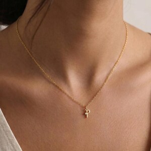 May include: A delicate gold-colored necklace with a small pendant in the shape of the letter "P". The necklace is worn around the neck. The background is a soft, neutral color.