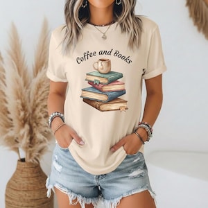 Könnte beinhalten: Cremefarbenes T-Shirt mit dem Text "Coffee and Books" über einem Stapel Bücher und einer Kaffeetasse. Die Bücher sind in Blau-, Rosa- und Brauntönen gehalten. Das Shirt hat kurze Ärmel.
