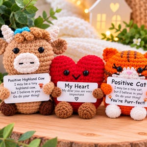 May include: Three crocheted plush toys: a tan Highland cow with a blue bow, a red heart, and an orange tabby cat. Each toy holds a white card with printed text. The cow card reads Positive Highland Cow, the heart card reads My Heart, and the cat card reads Positive Cat with the phrase You're Purr-fect.