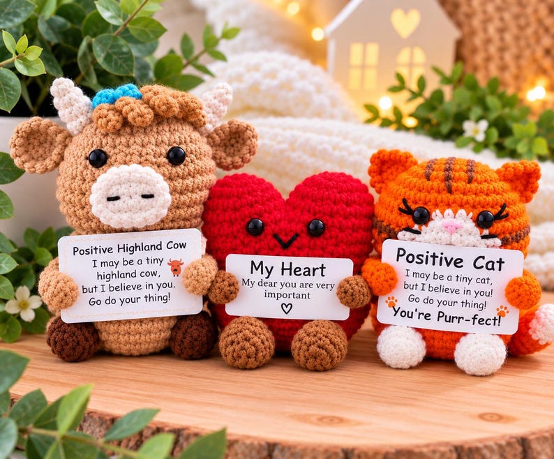 May include: Three crocheted plush toys: a tan Highland cow with a blue bow, a red heart, and an orange tabby cat. Each toy holds a white card with printed text. The cow card reads Positive Highland Cow, the heart card reads My Heart, and the cat card reads Positive Cat with the phrase You're Purr-fect.