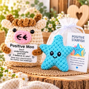 May include: A cream-colored crochet cow with brown hair and a pink snout holding a card that reads Positive Moo, alongside a bright blue crochet starfish and a card that reads Positive Starfish, both in a white organza gift bag.