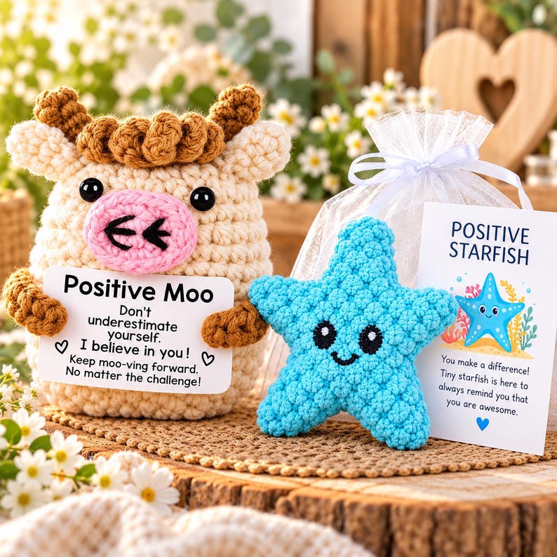 May include: A cream-colored crochet cow with brown hair and a pink snout holding a card that reads Positive Moo, alongside a bright blue crochet starfish and a card that reads Positive Starfish, both in a white organza gift bag.