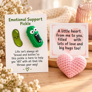 May include: A green textured pickle figurine with large eyes and an open mouth attached to a white card labeled Emotional Support Pickle, alongside a small pink crocheted heart and a square card with the text A little heart from me to you.