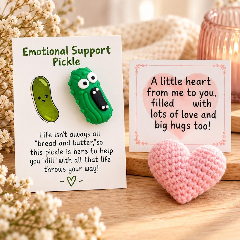 May include: A green textured pickle figurine with large eyes and an open mouth attached to a white card labeled Emotional Support Pickle, alongside a small pink crocheted heart and a square card with the text A little heart from me to you.