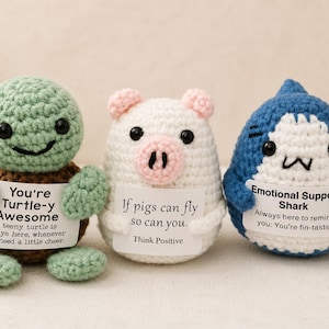 May include: Three crocheted plush figures: a green turtle, a white pig, and a blue shark. Each holds a white sign with text. The turtle sign reads: You're Turtle-y Awesome. The pig sign reads: If pigs can fly so can you. The shark sign reads: Emotional Support Shark.