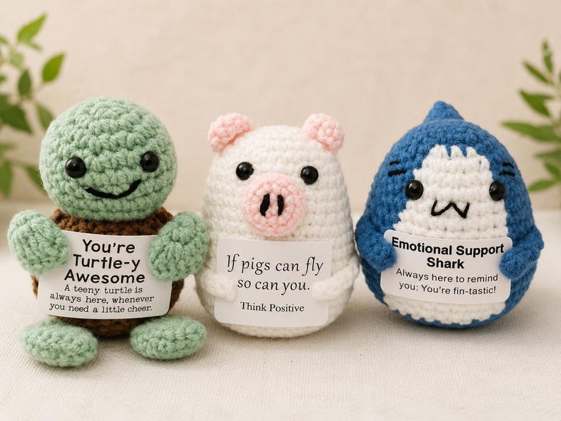 May include: Three crocheted plush figures: a green turtle, a white pig, and a blue shark. Each holds a white sign with text. The turtle sign reads: You're Turtle-y Awesome. The pig sign reads: If pigs can fly so can you. The shark sign reads: Emotional Support Shark.