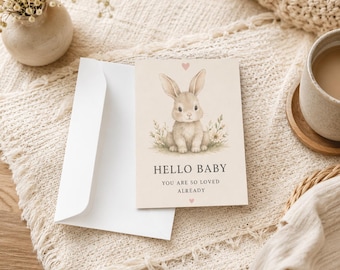 May include: A rectangular greeting card with a beige background featuring a watercolor illustration of a light brown bunny sitting in grass. Above the bunny is a small pink heart. The text reads HELLO BABY YOU ARE SO LOVED ALREADY.