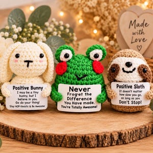 May include: Three small crocheted figures: a cream-colored bunny, a green frog with red cheeks, and a brown sloth. Each figure holds a white sign with black text featuring motivational messages including Positive Bunny, Never Froget, and Positive Sloth.