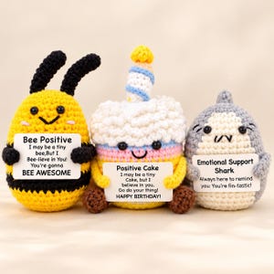 May include: Three crocheted plush toys: a yellow and black bee, a multi-colored birthday cake with a candle, and a gray shark. Each toy holds a white sign with printed text: Bee Positive, Positive Cake, and Emotional Support Shark.