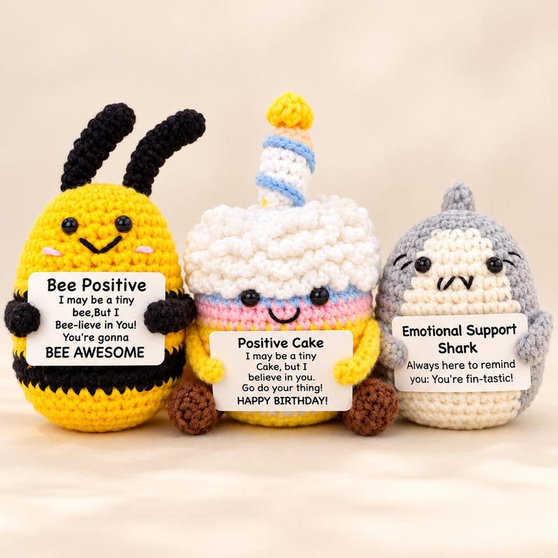 May include: Three crocheted plush toys: a yellow and black bee, a multi-colored birthday cake with a candle, and a gray shark. Each toy holds a white sign with printed text: Bee Positive, Positive Cake, and Emotional Support Shark.